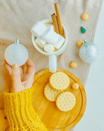 Beauty woman in yellow sweater holding Christmas ball for decoration with cacao marshmallow cinnamon, cookies and miniature Christmas tree on the tableの写真素材