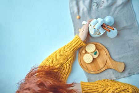 Pretty woman in a yellow sweater lying and drinking hot chocolate with marsmallows and cinnamon next to Christmas toys while decorating with cookies and miniature Christmas tree on the white tableの写真素材