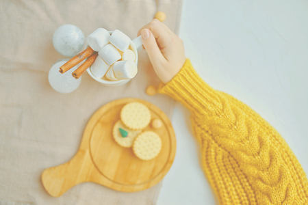 Cute woman in a yellow sweater drinking hot coffee with marsmallows and cinnamon next to Christmas toys while decorating with cookies and miniature Christmas tree on the white tableの写真素材