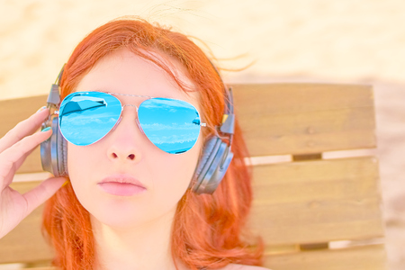 Beautiful woman listening to music on the beach. Portrait of a red-haired girl resting on the beach, lying on a deck chair and listening to relaxing music.の写真素材