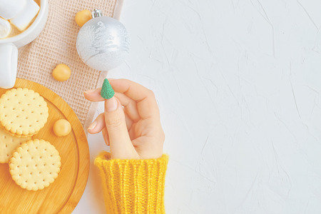 Beautiful woman in yellow sweater holding miniature Christmas tree with ornaments, hot coffee with marshmallow and cinnamon, cookies on the tableの写真素材