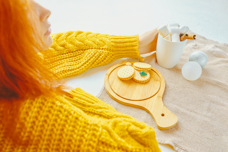 Beauty woman in a yellow sweater drinking hot chocolate with marsmallows and cinnamon next to Christmas toys while decorating with cookies and miniature Christmas tree on the white tableの写真素材