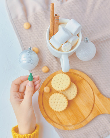 Cute woman in yellow sweater holding miniature Christmas tree with ornaments, hot cacao with marshmallow and cinnamon, cookies on the tableの写真素材