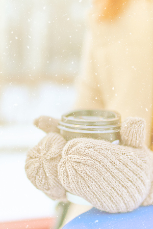 Awesome woman in a beige coat, yellow sweater, blue trousers, warm mittens drinks hot coffee to go in snowy weather in winterの写真素材
