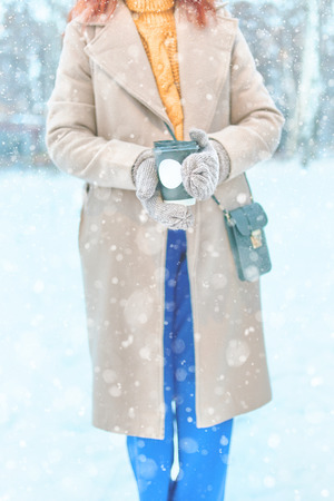 Red-haired woman in a beige coat, yellow sweater, blue trousers, warm mittens drinks hot coffee to go in snowy weather in winterの写真素材