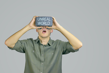 Young man in shirt enjoys and amazed by virtual reality glasses with "unreal world" inscription on them in front of light grey backgroundの写真素材