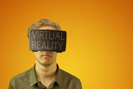 Young man in a shirt uses virtual reality glasses with the inscription "virtual reality" on them in front of a golden yellow backgroundの写真素材