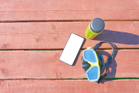 Swimming mask, bottle of smoothie and smartphone on wooden table on sunny day on vacation. mockupの写真素材
