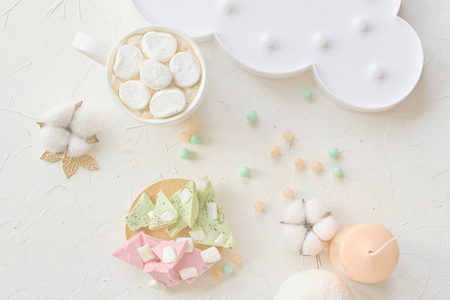 Hot coffee with marshmallow in a white mug next to lemon and strawberry chocolate, a cloud lamp, cotton and a candle on the tableの写真素材