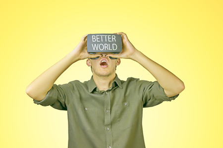Young man in shirt enjoys and amazed by virtual reality glasses with "better world" inscription on them in front of light yellow backgroundの写真素材