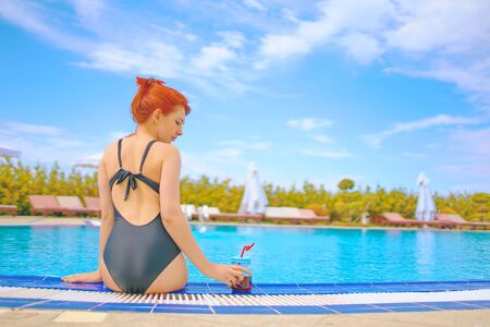 Cute woman sitting on the edge of the swimming pool holds smoothie mason jar in a tropical hotel on a Sunny dayの写真素材