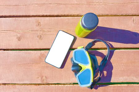 Swimming mask, bottle of smoothie and  smartphone on wooden table on sunny day on vacationの写真素材