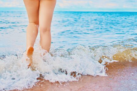 In the frame of a woman's feet walking on the beach by the sea on a Sunny day in a tropical hotel on vacation. Waves of ocean wash her feetの写真素材