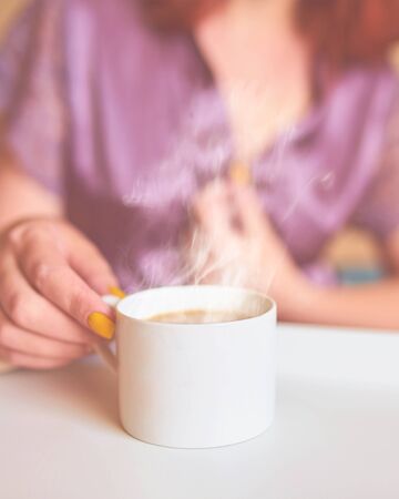 Cute woman in a purple silk robe drinking coffee early in the morning in bed. White cup of latte on the tray. Breakfast conceptの写真素材