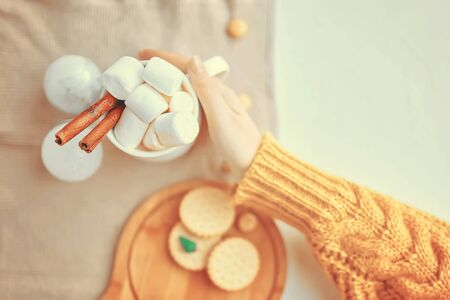 Woman in a yellow sweater drinks hot cocoa with marshmallow and cinnamon rolls in a white mug and cookies and a figure of a Christmas treeの写真素材