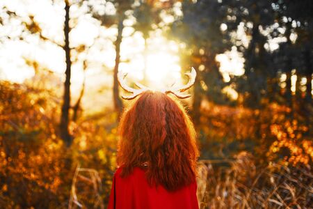 Woman in long red dress with deer horns in fall forest. Beautiful mystery gothic creature in autumn forest. Dark fantasy creative elf in magic woodの写真素材