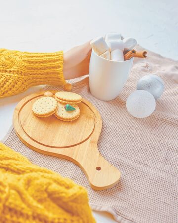 Woman in a yellow sweater drinks hot cocoa with marshmallow and cinnamon rolls in a white mug and cookies and a figure of a Christmas treeの写真素材