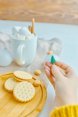 Woman in a yellow sweater drinks hot cocoa with marshmallow and cinnamon rolls in a white mug and cookies and a figure of a Christmas treeの写真素材