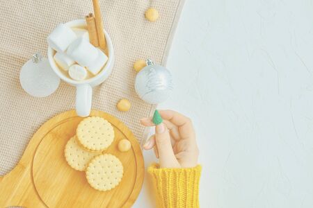 Woman in a yellow sweater drinks hot cocoa with marshmallow and cinnamon rolls in a white mug and cookies and a figure of a Christmas treeの写真素材