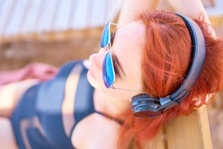 Cute woman listening to music on the beach. Red-haired young woman lying on the beach, relaxing on a lounger and listening to relaxing music.の写真素材