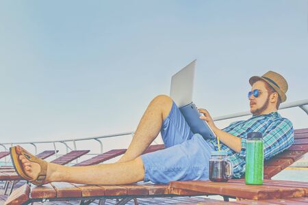 Young businessman resting on piers, enjoying summertime in resort spa near sea while working remotely at laptop computer connected to wireless internet.の写真素材