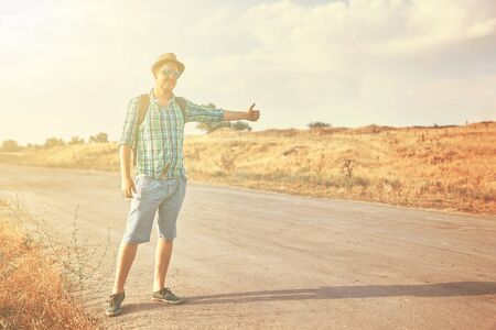 Hitchhiker with backpack in summer hat, light shirt, shorts on the road in tropical country in Sunny weather at autumnの写真素材