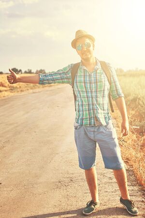 Hitchhiker with backpack in summer hat, light shirt, shorts on the road in tropical country in Sunny weather at autumnの写真素材
