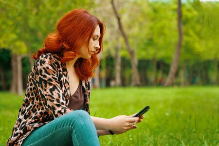 Young red haired woman in a leopard print coat and turquoise jeans is typing a message on her mobile phone while sitting on a lawn in a Parkの写真素材