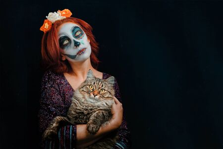 Young woman in day of the dead mask skull face art makeup and red hair with cat in hands, dreaming on dark background. Halloween conceptの写真素材