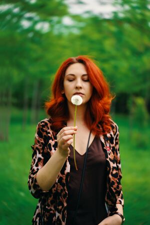 Walk in the Park. Portrait of a beautiful young laughing woman with red hair sitting on meadow with dandelion in hand, blurred background.の写真素材