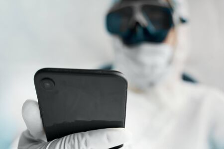 Doctor protects himself from a global pandemic and holds a dark phone in his hands to communicate with colleagues around the world about symptoms and the vaccineの写真素材
