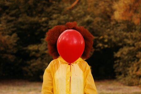 Red-haired curly afro woman in a yellow raincoat holding a red balloon on the background of forest....