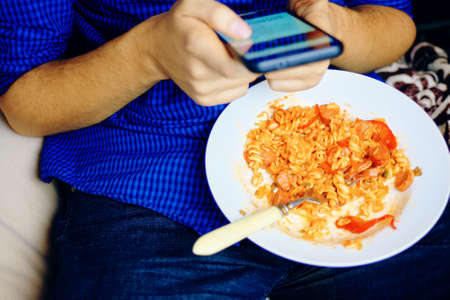 Man uses a smartphone in social networks while dining in a blue shirt and jeans sitting on a binbag during breakfast.の写真素材