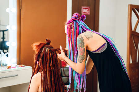Beautiful woman barber braids dreadlocks with tattoo making trendy braids dreadlocks in the her barbershop. Bishkek, Kyrgyzstan - July 04, 2019.のeditorial素材