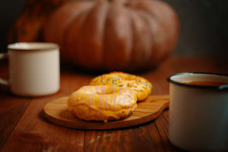 Autumn composition with metal mugs, donuts and pumpkin on a wooden background. Doughnuts with yellow lemon glaze and colorful sprinkles. Sweet pastries on a wooden tray.の写真素材