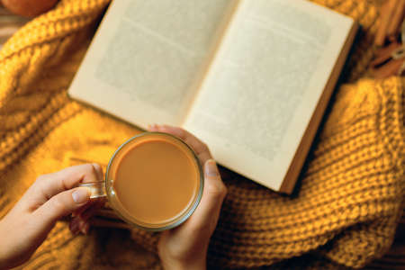 Cup of hot coffee in a womans hands and a book on orange knitted sweater. Cozy knitted warm orange sweater. Autumn-winter time. Open vintage book with romantic stories.の写真素材