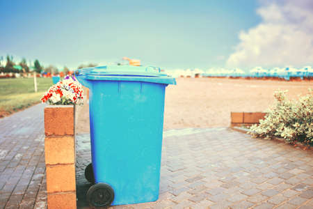Blue waste bucket on the shore of the lake. Trash can on the beach. Plastic large waste tank. Maintaining cleanliness in the recreation area of people. Beach with umbrellas and sun loungers.の写真素材