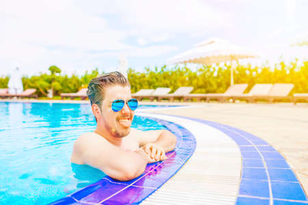 Man leans out of the pool, leaning on the edge. Close-up portrait of an attractive young man in sunglasses. Funny bearded man shows his tongue and smiles. Wooden deck chairs in front of green bushes.の写真素材