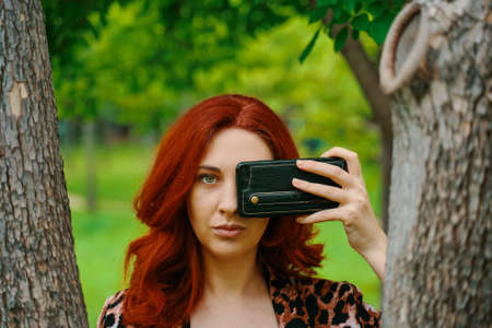 Girl in the Park. Red-haired woman takes a picture on her phone between the trees. Summer walk in the park. An example of excellent camera quality. Differences between phone camera and human eye.の写真素材