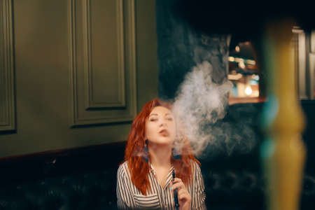 Pretty woman smoking hookah at the restaurant. Pleasure of shisha. Beautiful red-haired woman enjoys relaxing in bar. Young girl in striped shirt.の写真素材