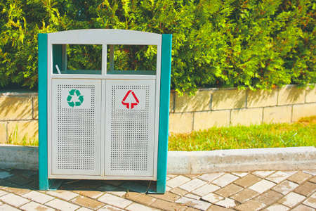 Trash can on the alley in the park area. Iron tank for different types of waste. Maintaining cleanliness in the recreation area of people. Green fir trees behind a brick fence.の写真素材