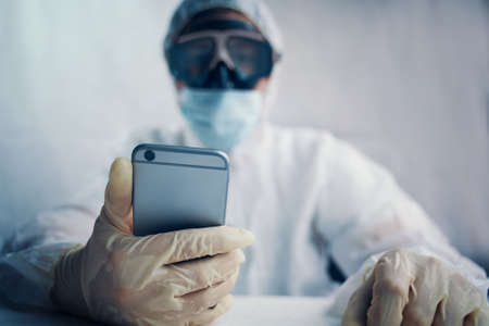 Virologist in a protective suit mask and gloves holds a phone in his hands for communication with family. Epidemic pandemic new rapidly spreading coronavirus 2019-ncov, medicine flu virus concept.の写真素材