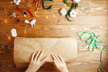 Top view of womens hands packing Christmas gift on wooden table. Wrapping craft paper with white dots design. Ribbons cotton boxes dry oranges garland and New Years toys on background.の写真素材