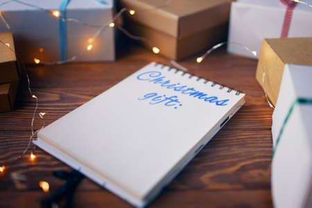 Wooden table with notepad with list of Christmas gifts garland and boxes. Concept of New Year. Space for writing. Boxes are tied with colored ribbons. White spiral notebook. Winter holiday.の写真素材