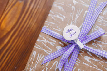 Craft paper with white abstract pattern. Gift box wrapped in paper with purple checkered ribbon on wooden table. Decorative clothespin with inscription Holly Jolly. Christmas eve. New Year mood.の写真素材