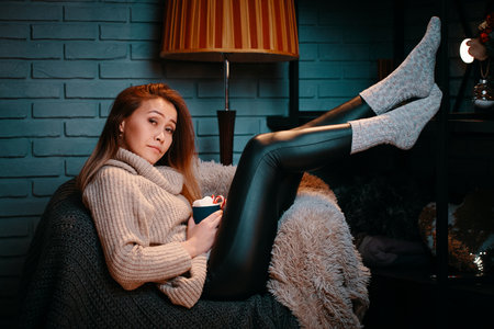 Woman with cup of cocoa on chair. Concept of Christmas and New year. Beautiful girl in sweater on knitted blanket. Gray brick wall on background. Winter vacation. Striped floor lamp behind chair.の写真素材