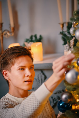 Cute guy hangs silver ball on Christmas tree. Festival decorations and bright garlands on fir. Fireplace with candles in background. Magic atmosphere. Winter holiday.の写真素材