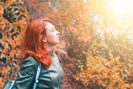 Young woman on the background of an fall forest. Portrait of a red-haired woman in the sun. A trip to nature. Autumn mountains. Fashionable womens jacket.の写真素材