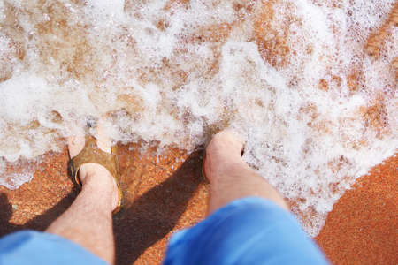 Top view of men's feet in sea. Man in flip-flops stands in the sand. An ocean wave washes to shore. Summer vacation.の写真素材