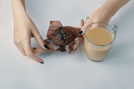 Womens hands unwrap chocolate muffin. Mug of coffee on white table. Morning romantic breakfast. Cocoa and delicious dessert. Copy space for advertising or marketing.の写真素材